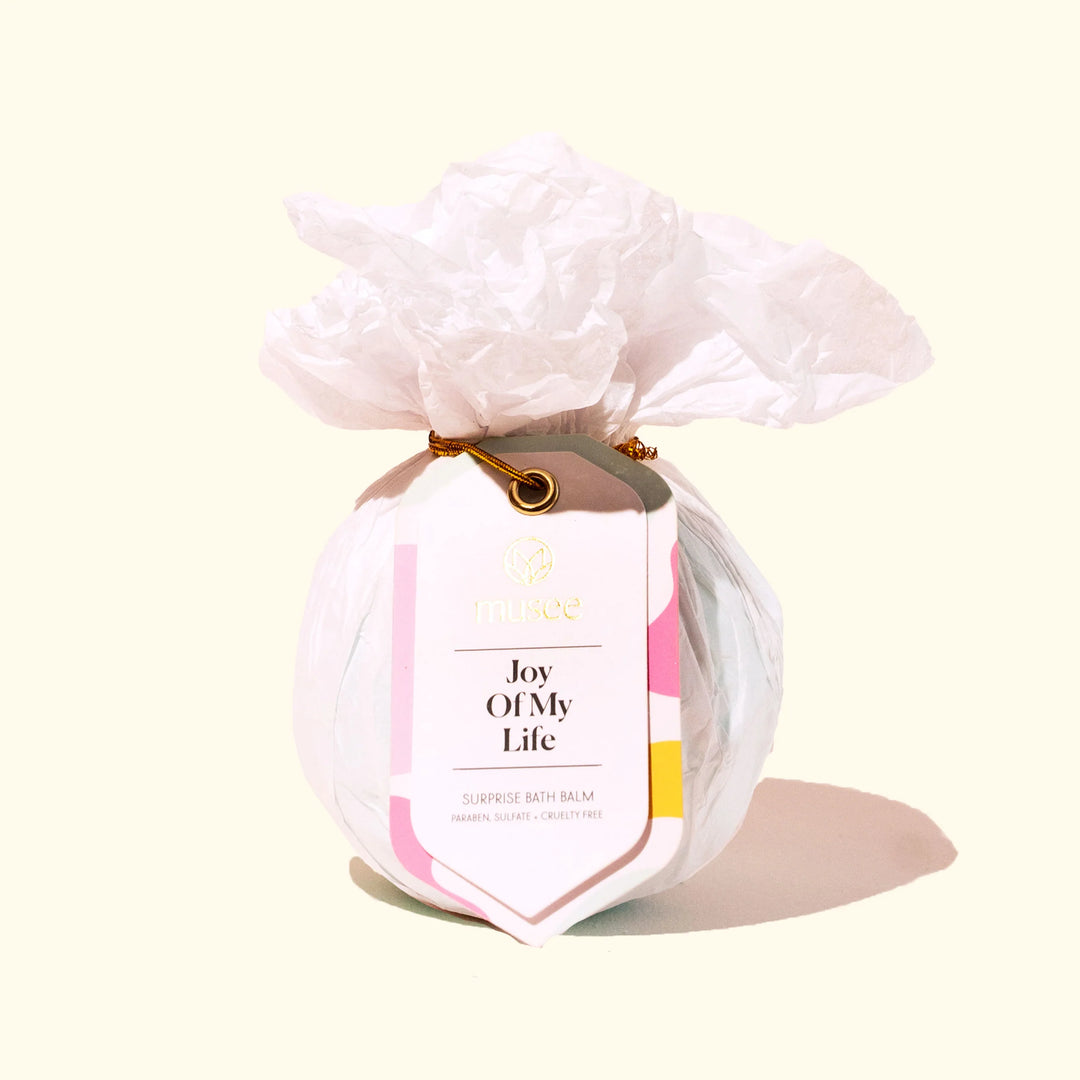Product photograph by Musee of a Joy Of My Life surprise bath balm wrapped in ruffled white tissue paper and secured with a gold cord and a tag featuring pink and yellow accents on a cream background