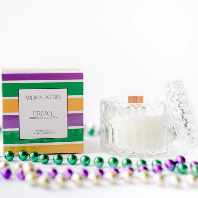 Commercial product photography by Aroma Avenue of a Krewe Mardi Gras Collection candle in a textured glass jar with a wooden wick and vibrant striped packaging surrounded by purple, green, and gold beads on a white background
