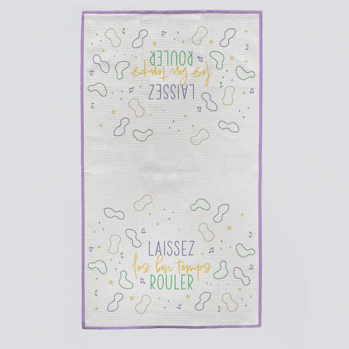 Illustrated fabric kitchen towel showcasing the iconic New Orleans Mardi Gras phrase Laissez les bon temps rouler in stylized lettering surrounded by whimsical peanut-shaped outlines, musical notes, and a purple stitched border