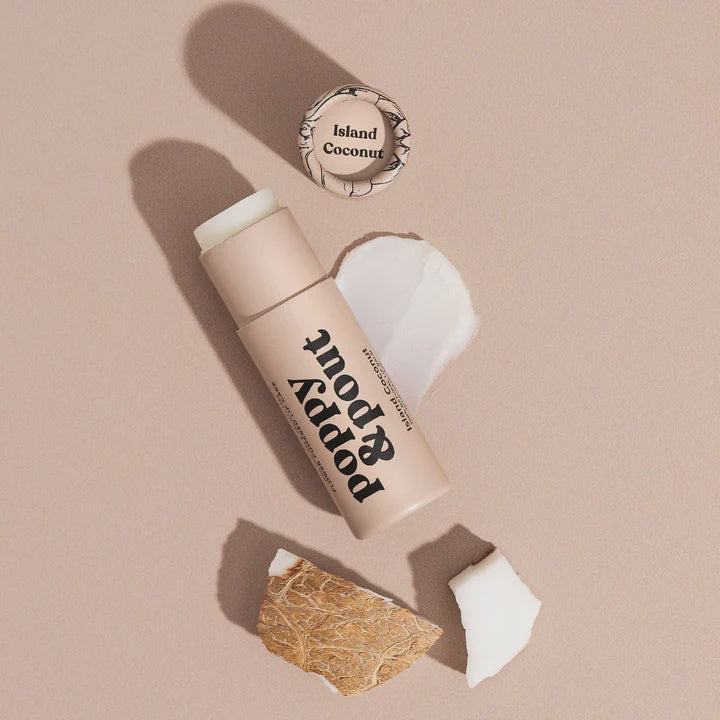 Product photograph by Poppy &amp; Pout of an Island Coconut lip balm on a neutral beige surface featuring the brand logo, a creamy white balm smear, and chunks of textured brown coconut husk
