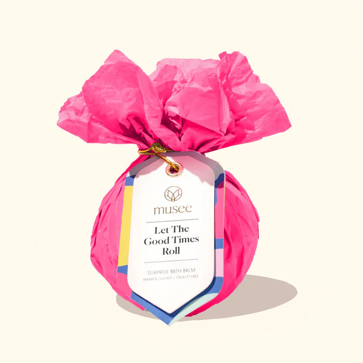 Product photograph by Musee of a surprise bath balm wrapped in vibrant pink crinkled tissue paper featuring a white decorative tag with Let The Good Times Roll text and a gold wire tie against a plain cream background