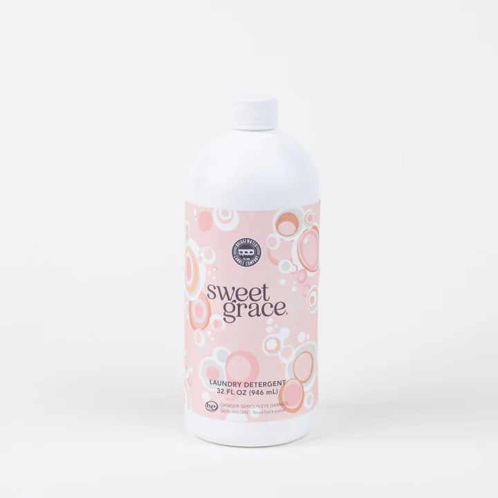 Product photography by Bridgewater Candle Company of a 32-ounce white bottle of Sweet Grace laundry detergent in a studio setting featuring a pink label with circular bubble patterns and elegant serif typography