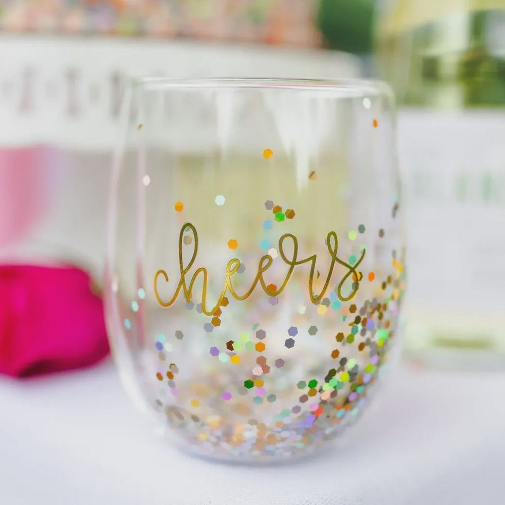 Photograph of a celebratory stemless wine glass featuring gold cursive cheers text and vibrant multicolored hexagonal glitter on a white tabletop with a blurred pink floral background