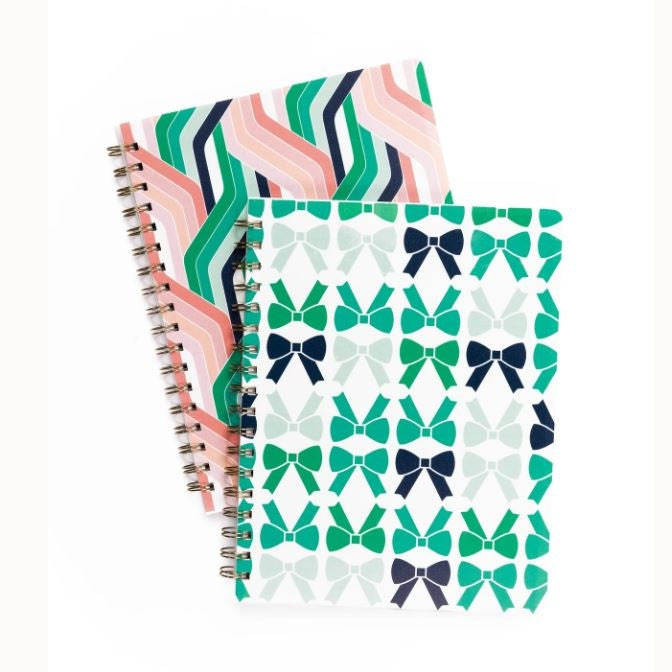 Two spiral notebooks by Lilly Pulitzer featuring a geometric chevron pattern in coral and emerald and a repeating bow motif in navy and teal on a clean white background with gold-tone wire-o binding