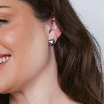 Photograph of a woman wearing a silver pyramid stud earring in a close-up studio shot featuring her long brown hair and a cheerful smile