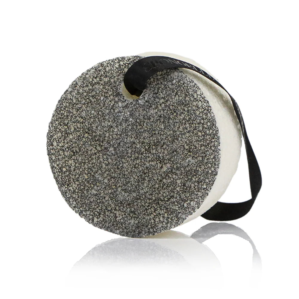 Product photograph by Spongellé of a circular Body Wash Infused Buffer on a white reflective surface featuring a textured grey speckled exfoliating side and a black hanging ribbon with the brand logo