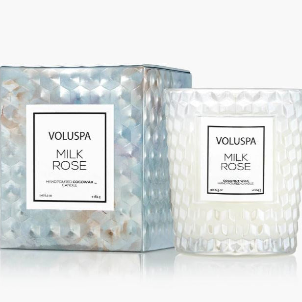 Product photograph by Voluspa of a Milk Rose candle and box featuring an iridescent pearlescent finish with a raised geometric honeycomb texture and minimalist white square labels on a reflective white surface