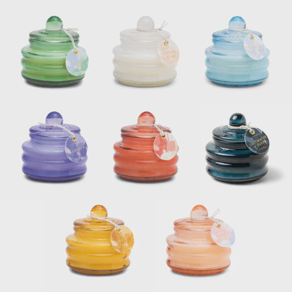 Product photography of eight Capri Blue beehive-style glass candles in a studio setting, featuring a vibrant spectrum of colors including emerald green and lavender with circular paper tags tied by delicate string