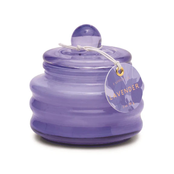 Product photography of a Paddywax lavender candle in a translucent purple ribbed glass jar featuring a matching spherical knob lid and a circular paper tag with gold lettering tied with a white string against a white background