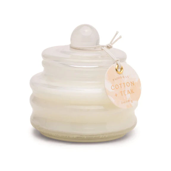 Scented candle by Paddywax featuring white Cotton and Teak wax in a tiered glass jar on a white background with a spherical lid handle and a peach watercolor tag tied with a string