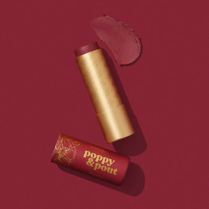 Commercial photograph for Poppy &amp; Pout featuring a Penny lip tint and floral cap on a burgundy background with a textured product smear, repeating gold foil logos, and eco-friendly cardboard packaging