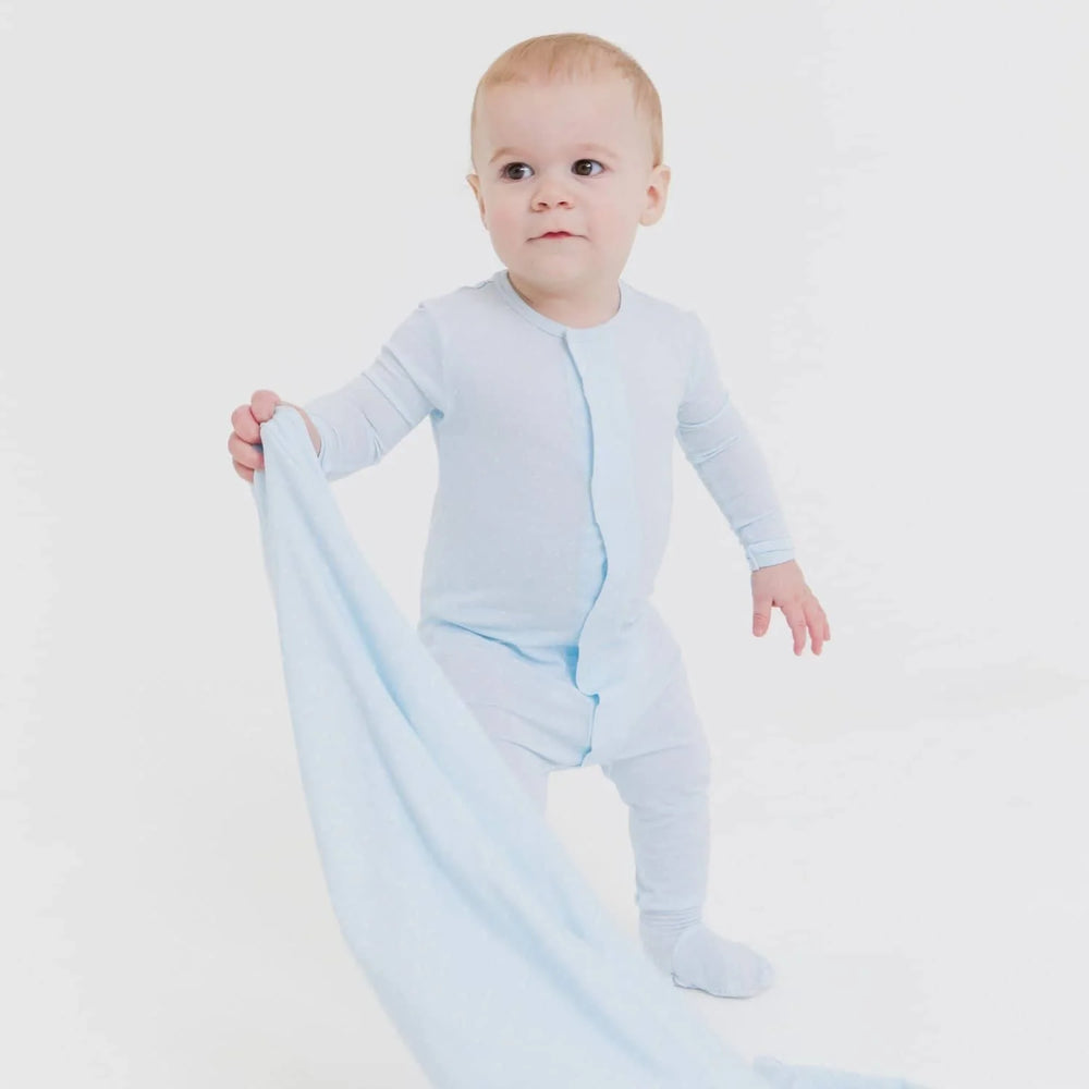 Studio photograph of an infant boy standing in a minimalist white studio wearing a light blue zippered footie romper while holding a matching soft blue security blanket with a curious expression
