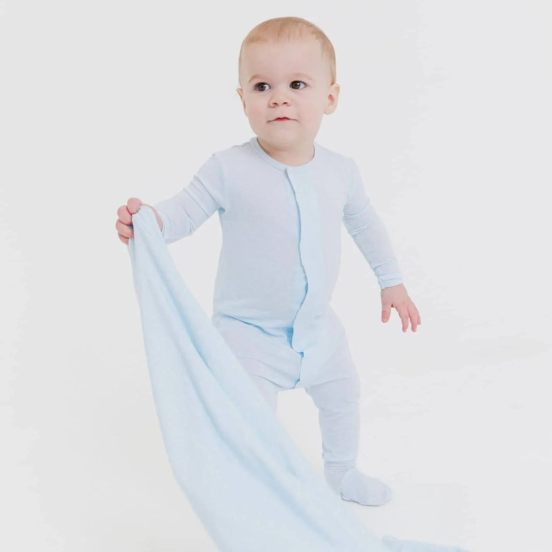 Studio photograph of an infant boy standing in a minimalist white studio wearing a light blue zippered footie romper while holding a matching soft blue security blanket with a curious expression