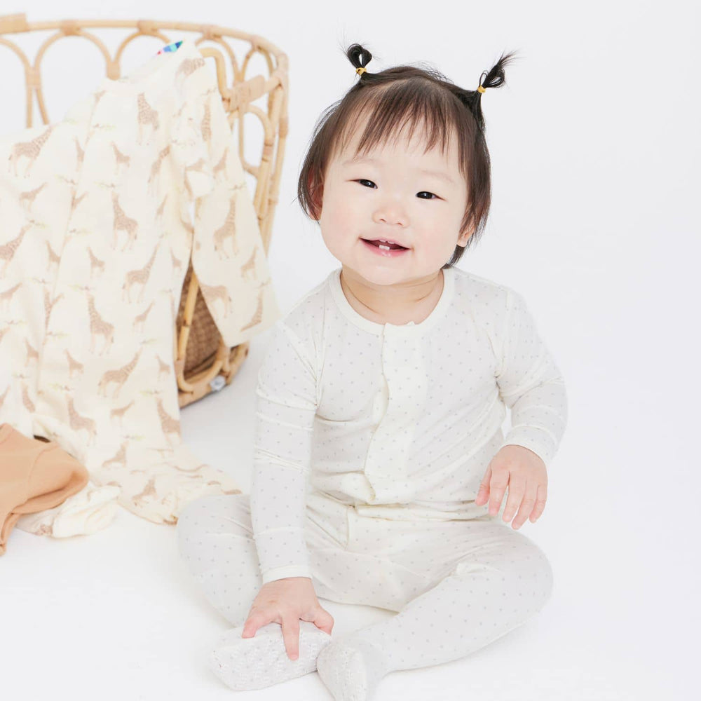 Digital photograph of a smiling East Asian baby girl in a bright white studio featuring two tiny top-knot pigtails, cream-colored dotted pajamas, and a giraffe-patterned blanket draped over a woven rattan basket