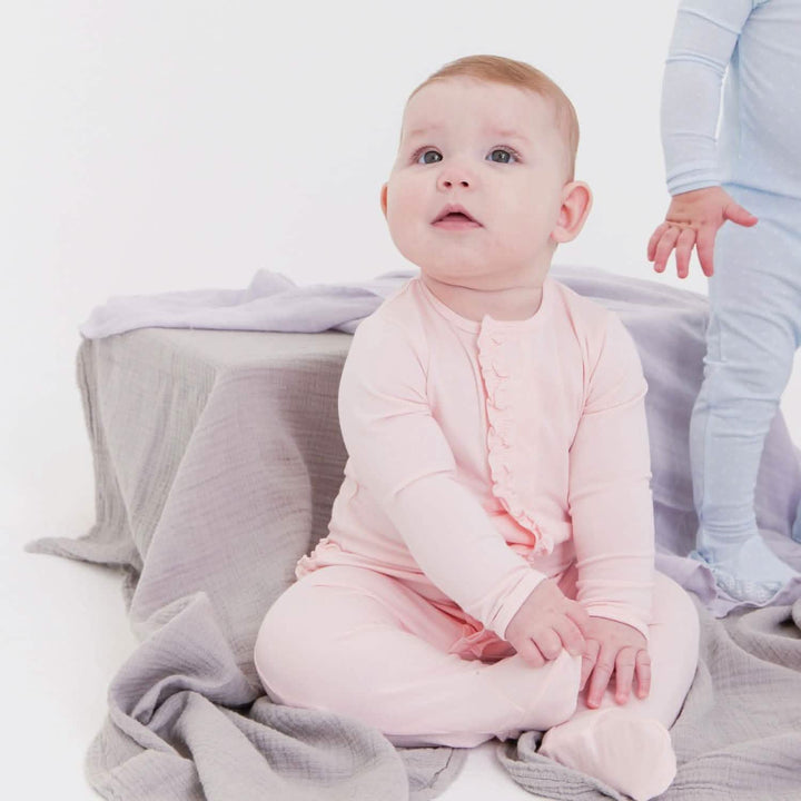 Photograph of a baby sitting in a minimalist white studio wearing a light pink ruffled romper and looking upward beside draped grey muslin cloth