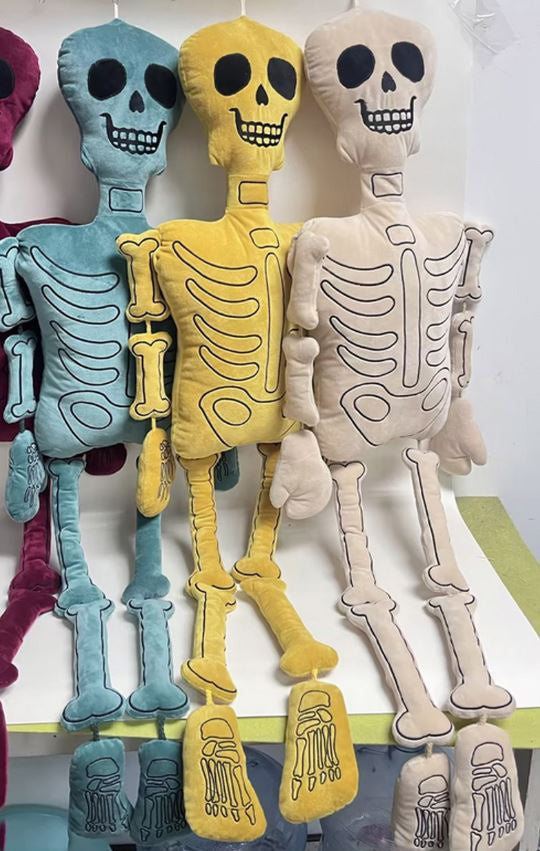 Plush fabric skeleton dolls in teal, yellow, and beige hanging in a studio setting featuring black embroidered bone outlines, hollow eye sockets, and long segmented limbs
