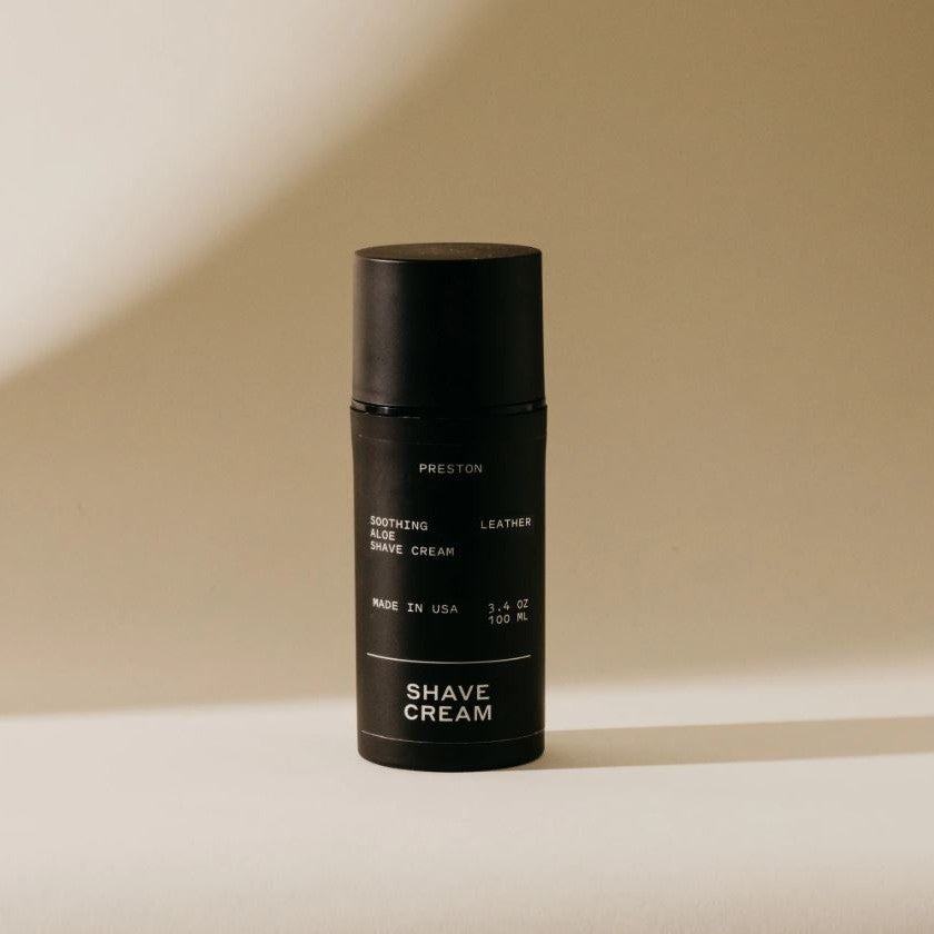 Studio photograph of a matte black Preston Soothing Aloe Shave Cream bottle with a leather scent label featuring minimalist white typography and a soft directional shadow on a beige studio backdrop