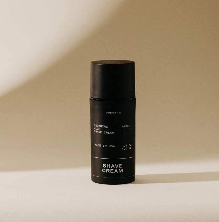 Studio photograph of a Preston soothing aloe shave cream bottle in a minimalist beige studio featuring a matte black finish, white sans-serif typography, and a soft shadow against a curved background