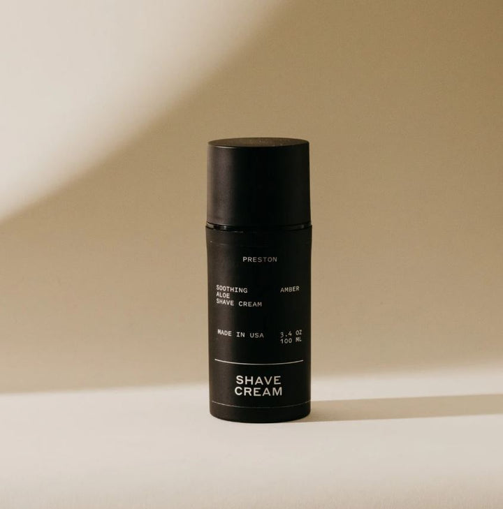 Studio photograph of a Preston soothing aloe shave cream bottle in a minimalist beige studio featuring a matte black finish, white sans-serif typography, and a soft shadow against a curved background