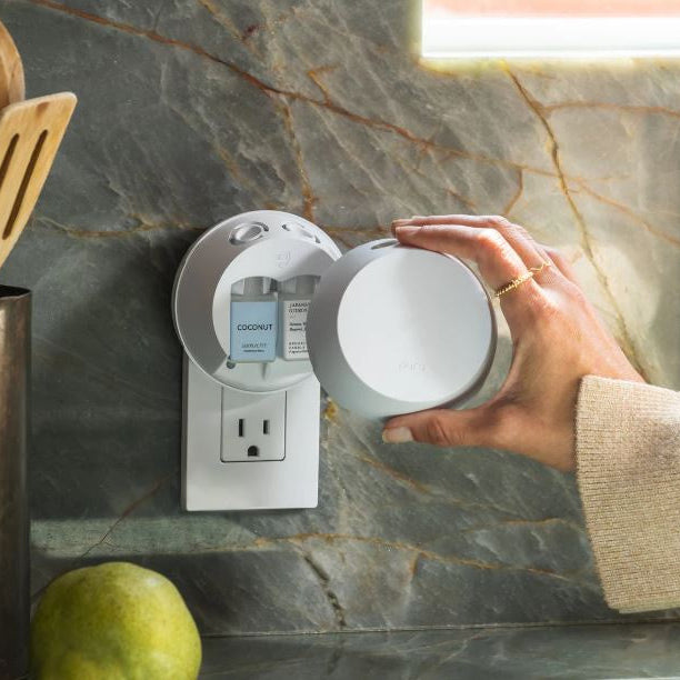 Digital photograph by Pura of a smart home fragrance diffuser plugged into a wall outlet against a marble backsplash with a hand wearing a gold ring inserting a Coconut scented vial next to a fresh green pear