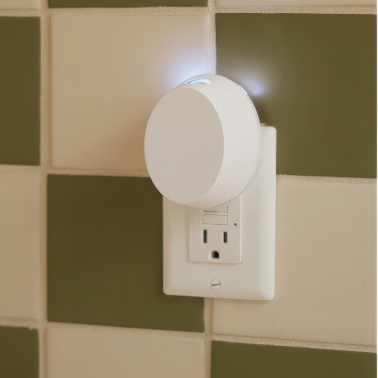 Digital photograph of a Pura smart fragrance diffuser plugged into a white wall outlet against olive green and cream checkered tiles featuring a soft blue indicator light at the top rim