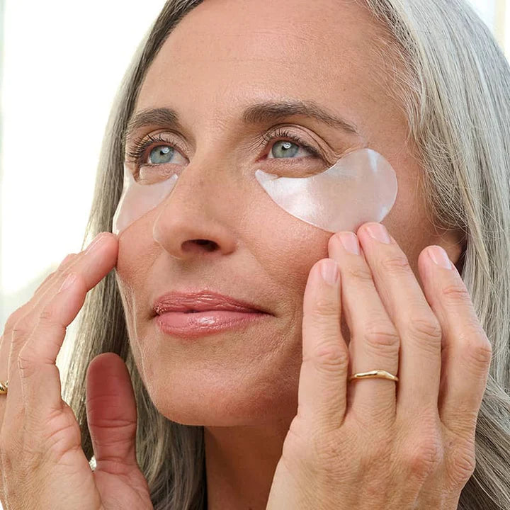 Digital photograph of a mature woman with long grey hair in a bright indoor setting wearing translucent shimmering gel patches under her blue eyes and a slim gold ring