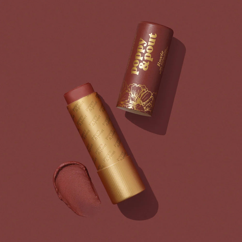 Commercial studio photograph for Poppy &amp; Pout of a Roxie tinted lip balm tube on a dark red background featuring gold foil lettering, an intricate floral design, and a creamy product swatch