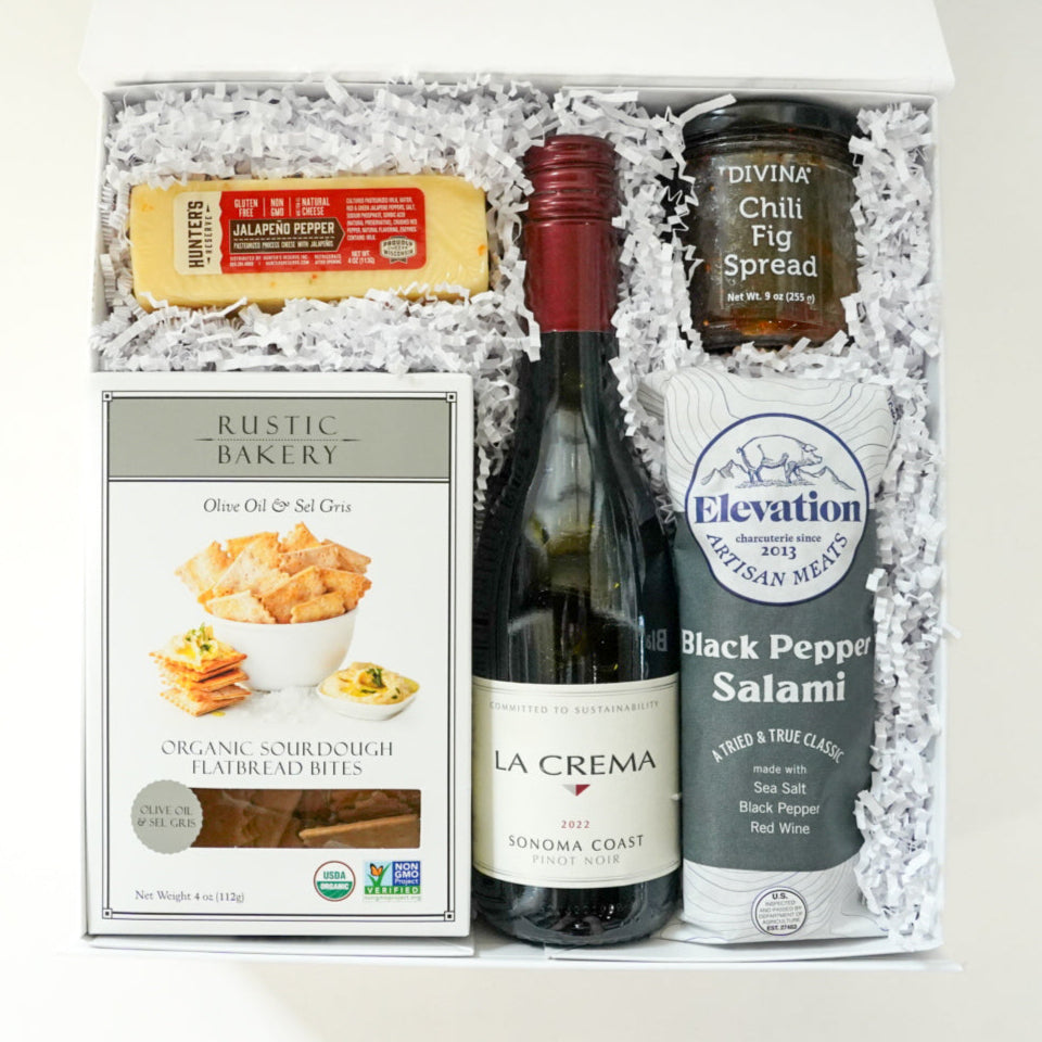Photograph of a curated gourmet gift box featuring a red-capped bottle of La Crema Pinot Noir, a bright yellow block of jalapeño cheese, and Elevation Meats black pepper salami nestled in white crinkle paper