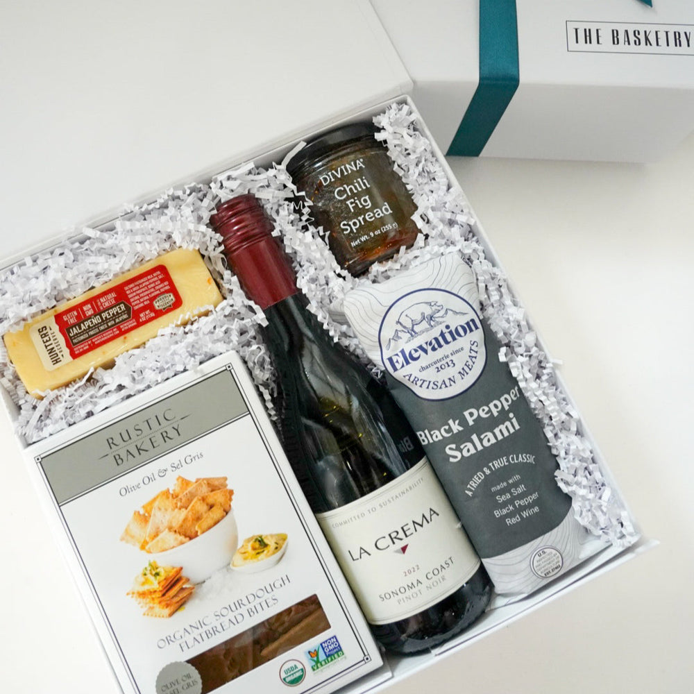 Photograph by The Basketry of a gourmet charcuterie gift set featuring a bottle of La Crema Pinot Noir, black pepper salami, and a bright yellow block of jalapeño pepper cheese nestled in white crinkle paper with The Basketry brand logo visible on the gift box lid