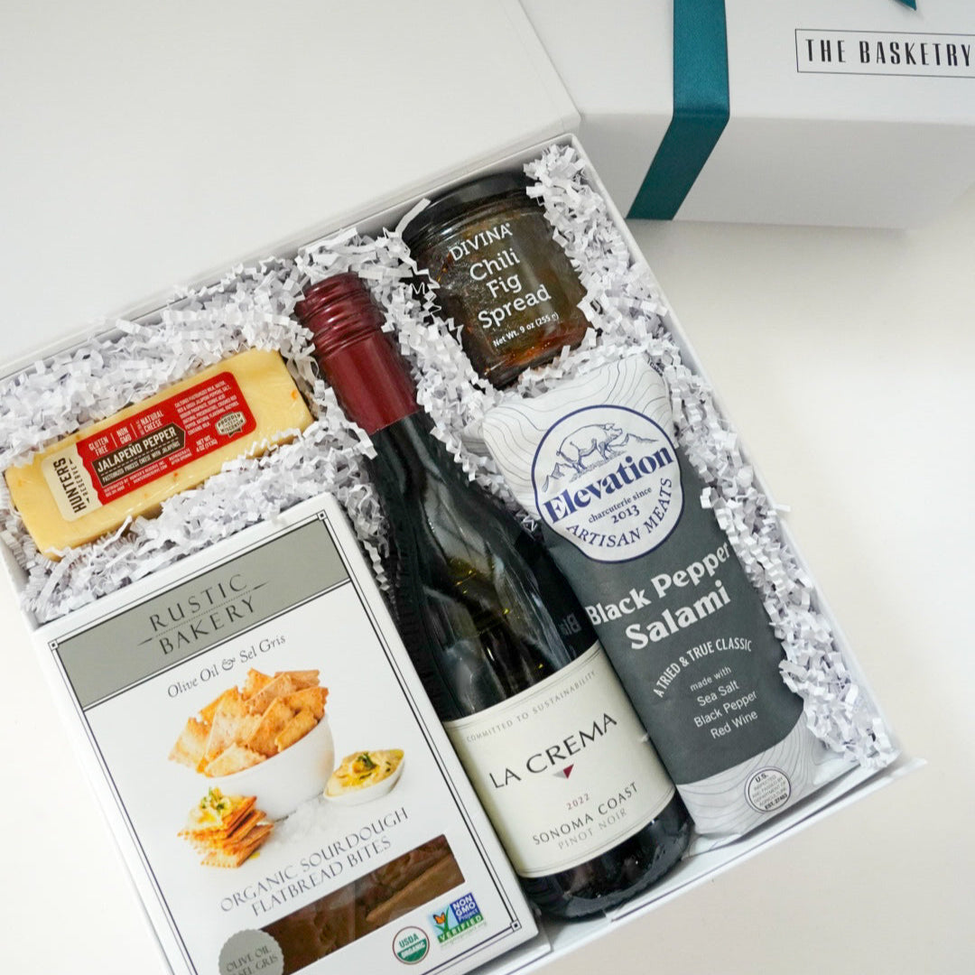 Photograph by The Basketry of a gourmet charcuterie gift set featuring a bottle of La Crema Pinot Noir, black pepper salami, and a bright yellow block of jalapeño pepper cheese nestled in white crinkle paper with The Basketry brand logo visible on the gift box lid