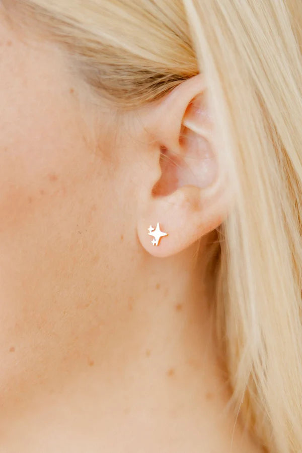 Photograph of a gold North Star sparkle stud earring worn on a person's earlobe featuring a dainty four-pointed star design framed by soft blonde hair and fair skin