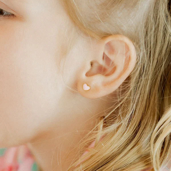Digital photograph of a young girl's ear in a close-up lifestyle shot featuring a small pale pink heart-shaped stud earring nestled among strands of wispy blonde hair and soft sunlit skin