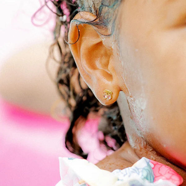 Photograph of a child's profile at a sunny location showcasing a gold cherry-shaped earring with pink stones, wet dark curls, and white sunscreen on the skin against a vibrant pink background