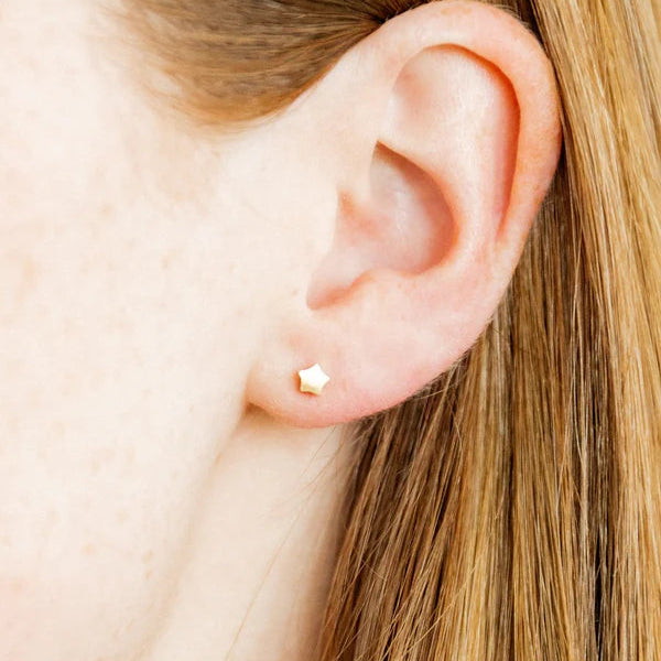 Photograph of a minimalist gold star stud earring positioned on the lower earlobe, detailing the helix and tragus curves alongside light freckles and strands of straight golden hair tucked behind the ear