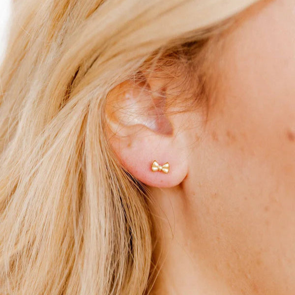 Digital photograph of a dainty yellow gold bow stud earring worn on a woman's earlobe featuring a tiny polished ribbon design and soft strands of blonde hair