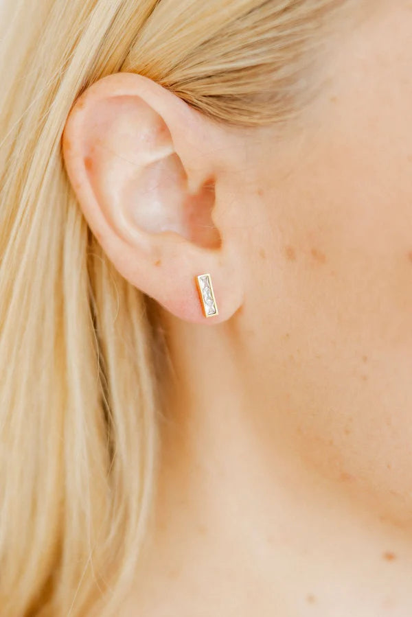 Photograph of a woman's earlobe showcasing a rectangular gold bar earring set with three small clear stones, framed by blonde hair and fair skin with subtle freckles