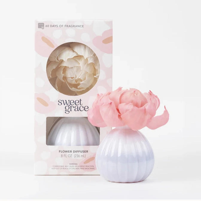Studio product photograph of a Sweet Grace flower diffuser by Bridgewater Candle Company featuring a fluted lavender glass vase with a large pink blooming flower and its pastel floral-patterned packaging on a plain white background