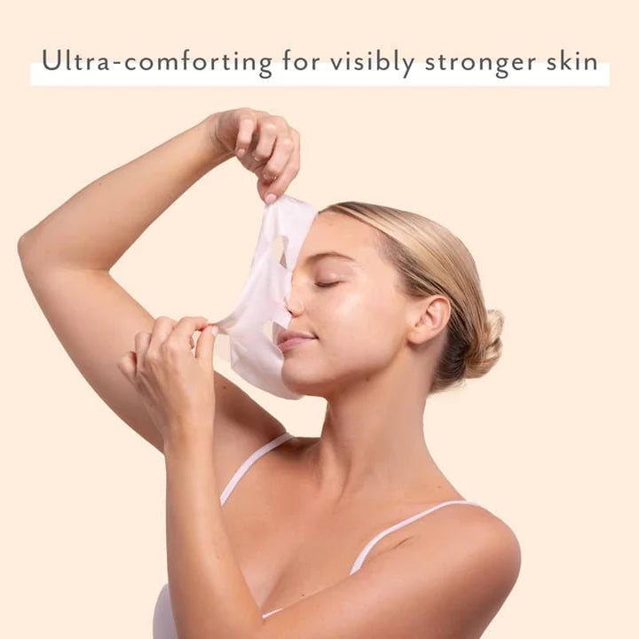 Commercial photograph of a woman in a studio setting peeling a white sheet mask from her face featuring blonde hair in a bun and a white spaghetti-strap top against a neutral beige background with text that says Ultra-comforting for visibly stronger skin
