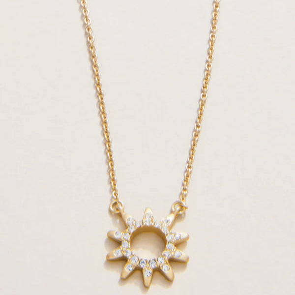 Gold jewelry necklace featuring a sunburst pendant centered on a plain off-white background with shimmering crystals pavé-set into the sun rays and a delicate gold cable chain