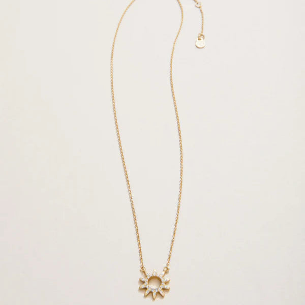 Jewelry photograph of a gold sunburst necklace featuring a hollow-center pendant with shimmering clear pavé stones and a delicate adjustable cable chain on a neutral off-white background