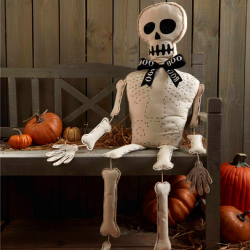 Photograph of a plush white fabric skeleton sitting on a grey wooden bench surrounded by orange pumpkins and straw featuring a black BOO ribbon and dashed stitching on its bones