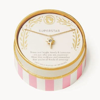 Studio photograph of an 18kt gold-plated starburst necklace with a central crystal stone showcased in a pink and white striped round gift box featuring gold foil Superstar lettering and laurel wreath illustrations on a white background