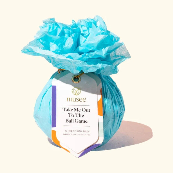 Commercial product photography of a Musee Take Me Out To The Ball Game surprise bath balm wrapped in vibrant blue crinkled tissue paper featuring a white tag with a gold eyelet and colorful geometric accents on a cream background
