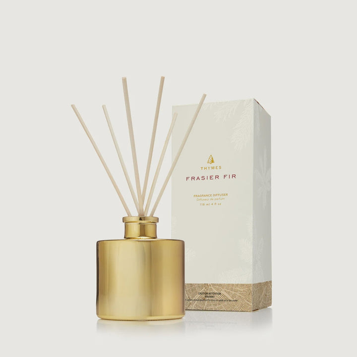 Studio photograph by Thymes of a Frasier Fir Fragrance Diffuser on a neutral background featuring a reflective gold metallic bottle, fanned light wood reeds, and the iconic Thymes logo on a white pine-patterned box