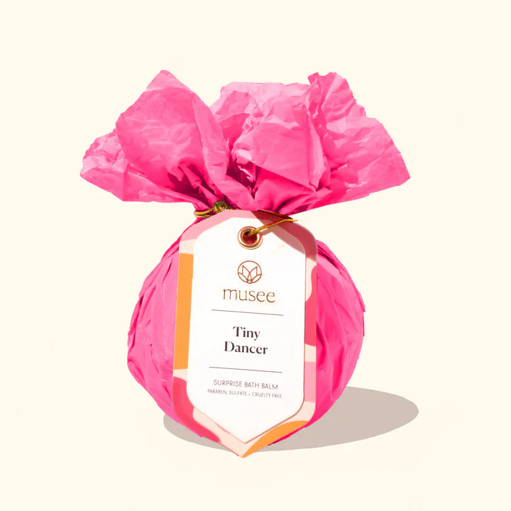 Studio product photograph by Musee Bath of a Tiny Dancer surprise bath balm wrapped in vibrant pink crinkled tissue paper tied with a gold metallic cord and featuring a white logo tag on a neutral cream background