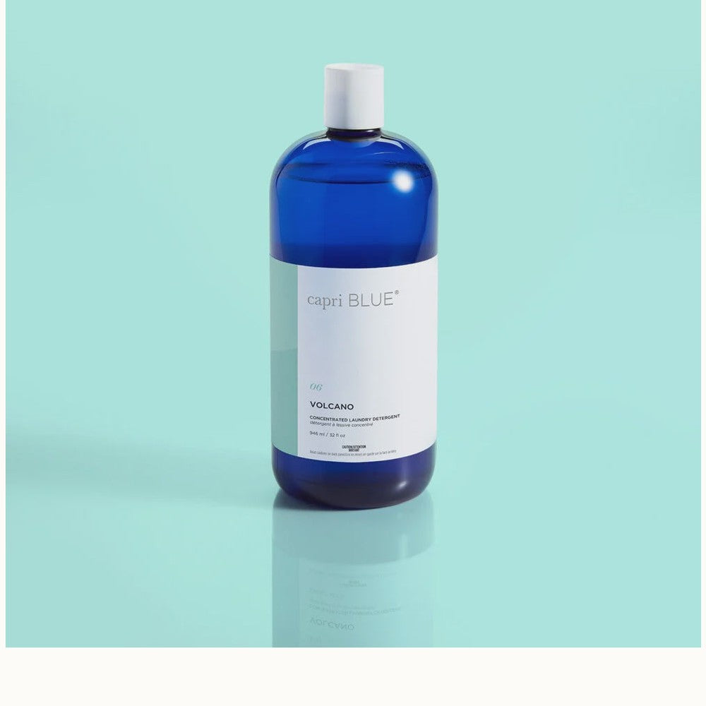 Studio photograph of Capri Blue Volcano Concentrated Laundry Detergent featuring a translucent cobalt blue bottle with a minimalist white label and white cap centered on a reflective surface against a solid light teal background