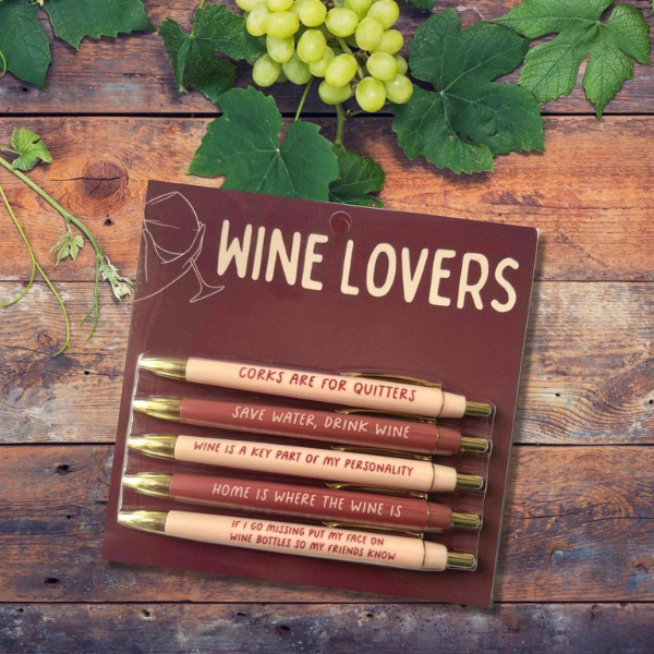 Photograph of a Wine Lovers pen set featuring five burgundy and cream gold-tipped pens with humorous wine slogans displayed on a rustic wood surface next to fresh green grapes and lush vine leaves