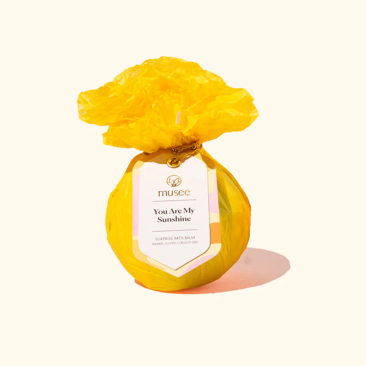 Product photograph by Musee of a You Are My Sunshine surprise bath balm wrapped in vibrant yellow crinkled paper with a decorative ruffled top and a gold-tied label on a cream background