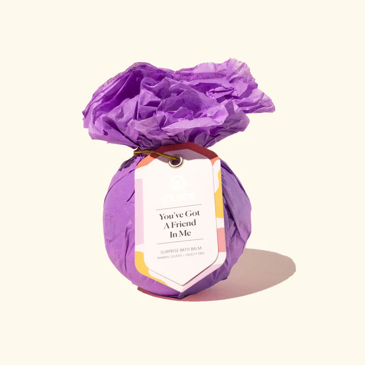 Studio photograph of a Musee surprise bath balm wrapped in vibrant purple crinkled tissue paper featuring a gift tag with the text You&