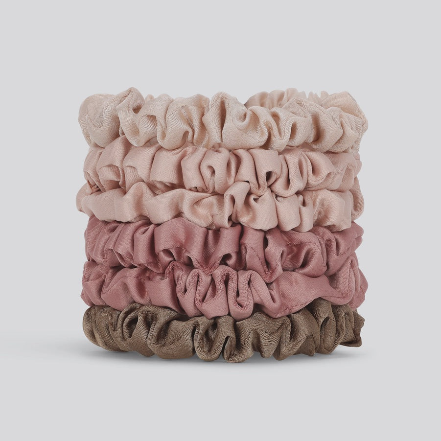 Studio product photograph of four silk satin scrunchies stacked vertically in a gradient of cream, dusty rose, and taupe shades against a neutral background showcasing ruffled textures and a soft lustrous sheen